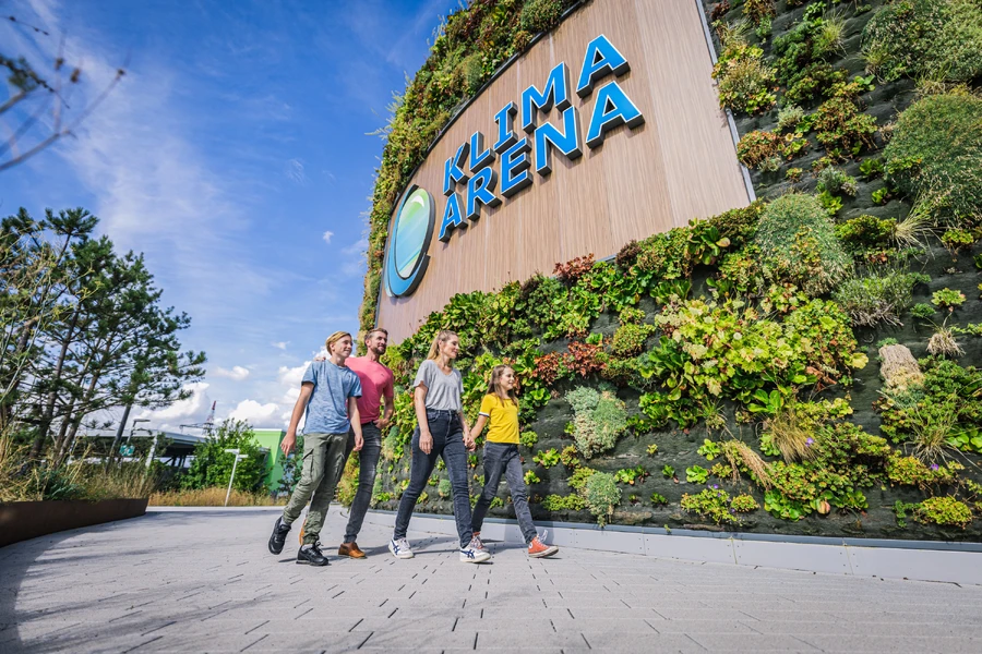Familie l&auml;uft auf dem Vorplatz der KLIMA ARENA entlang der begr&uuml;nten Fassade mit Logo.